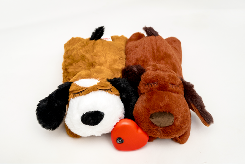 Two plush dog toys resembling a puppy and a brown dog with a heartbeat device inside, alongside a red bell.