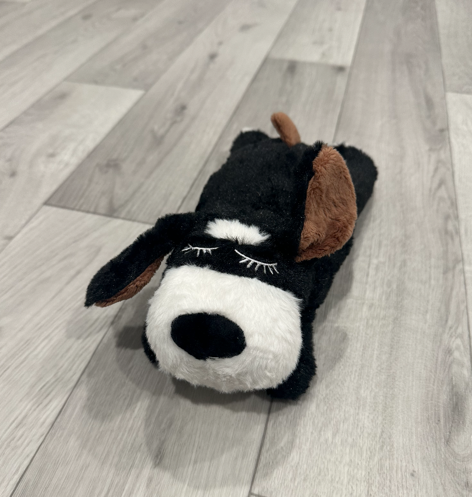 Black and white plush dog toy with brown ears and stitched eyelids, lying on a gray wood floor, designed for cuddling and light play.