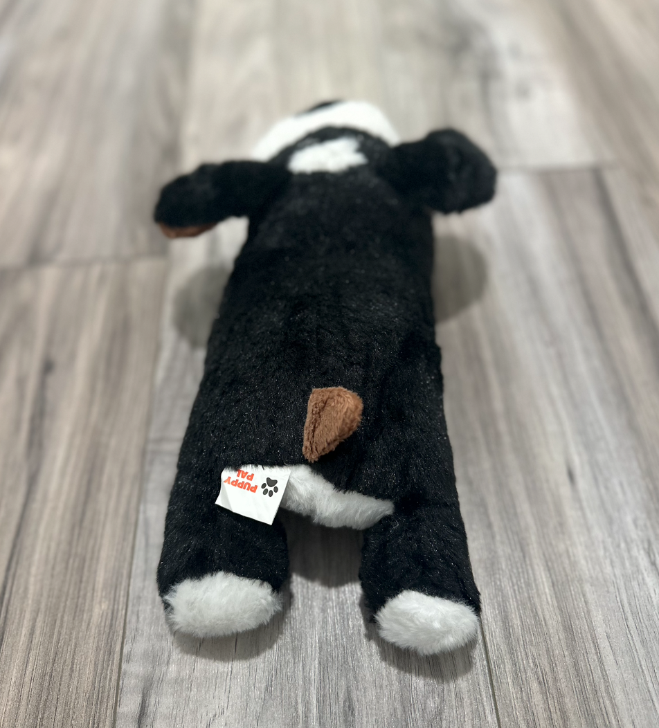 Back view of a black and white plush dog toy with brown tail and ears, lying on a wood floor with visible product tag.